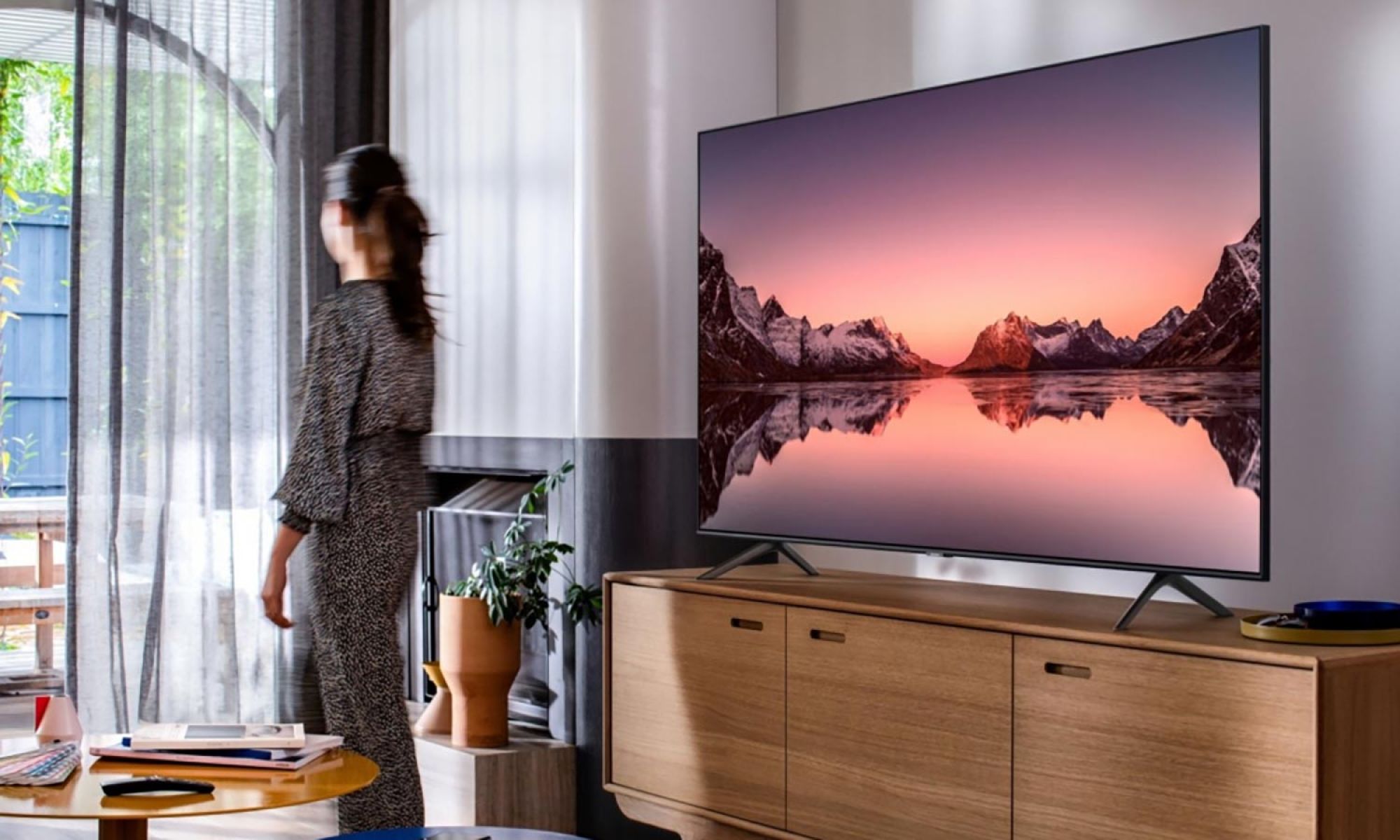 A woman looks out the window in a living room with a Samsung large flat-screen TV displaying a scenic landscape.