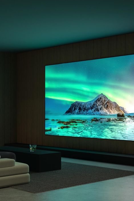 A home theater with a large screen showing a scenic view of a mountain and aurora borealis over the ocean, with a modern sofa in the foreground.