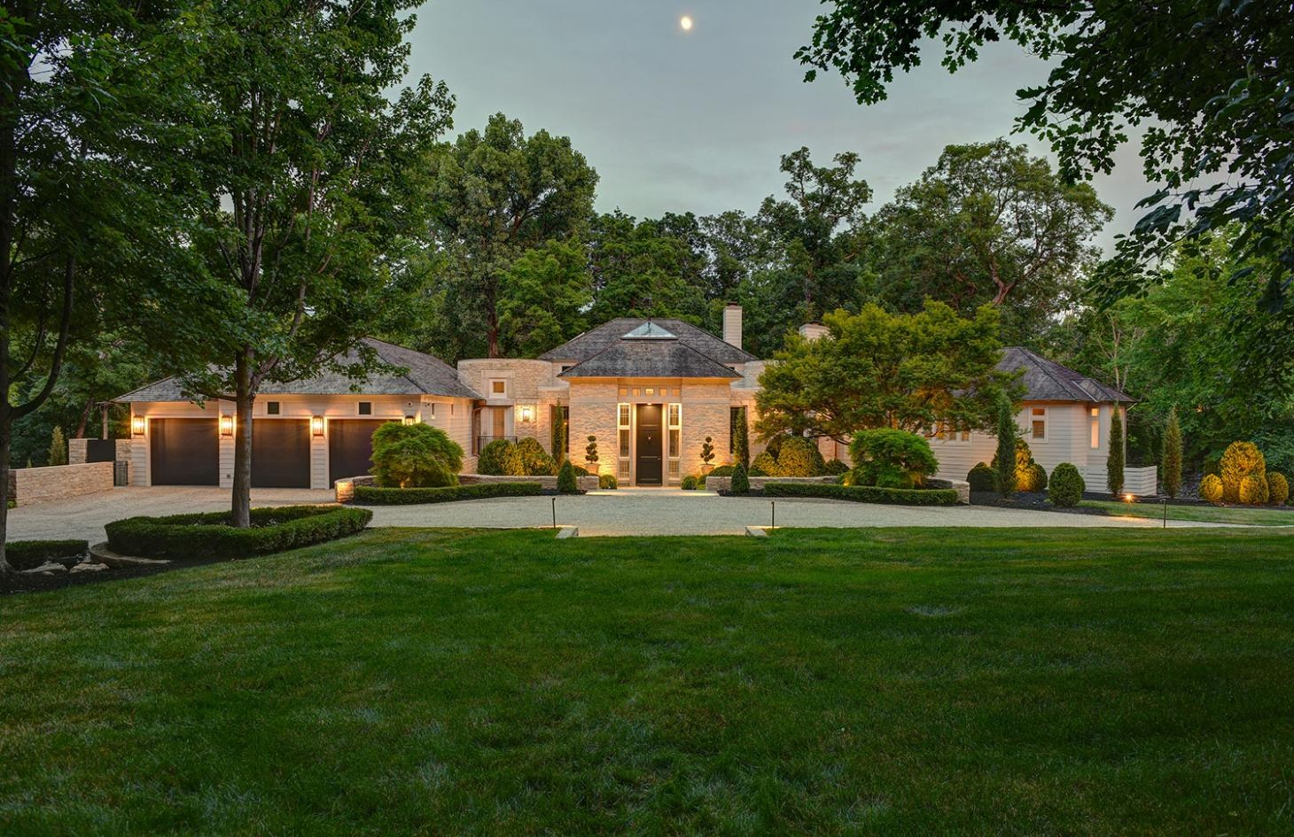 A luxurious home exterior at dusk, featuring a well-lit entrance, manicured landscaping, and a three-car garage framed by trees.