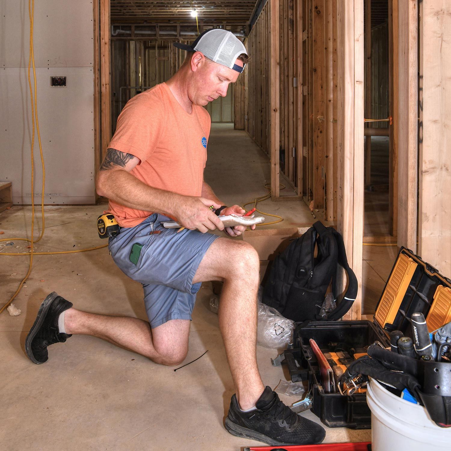 Electrician kneeling on the floor while working with wiring in a framed house under construction.