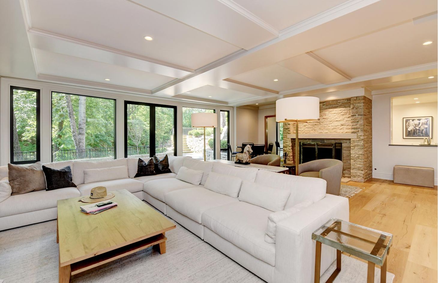 A cozy family room with a large white sectional, stone fireplace, and floor-to-ceiling windows overlooking greenery.