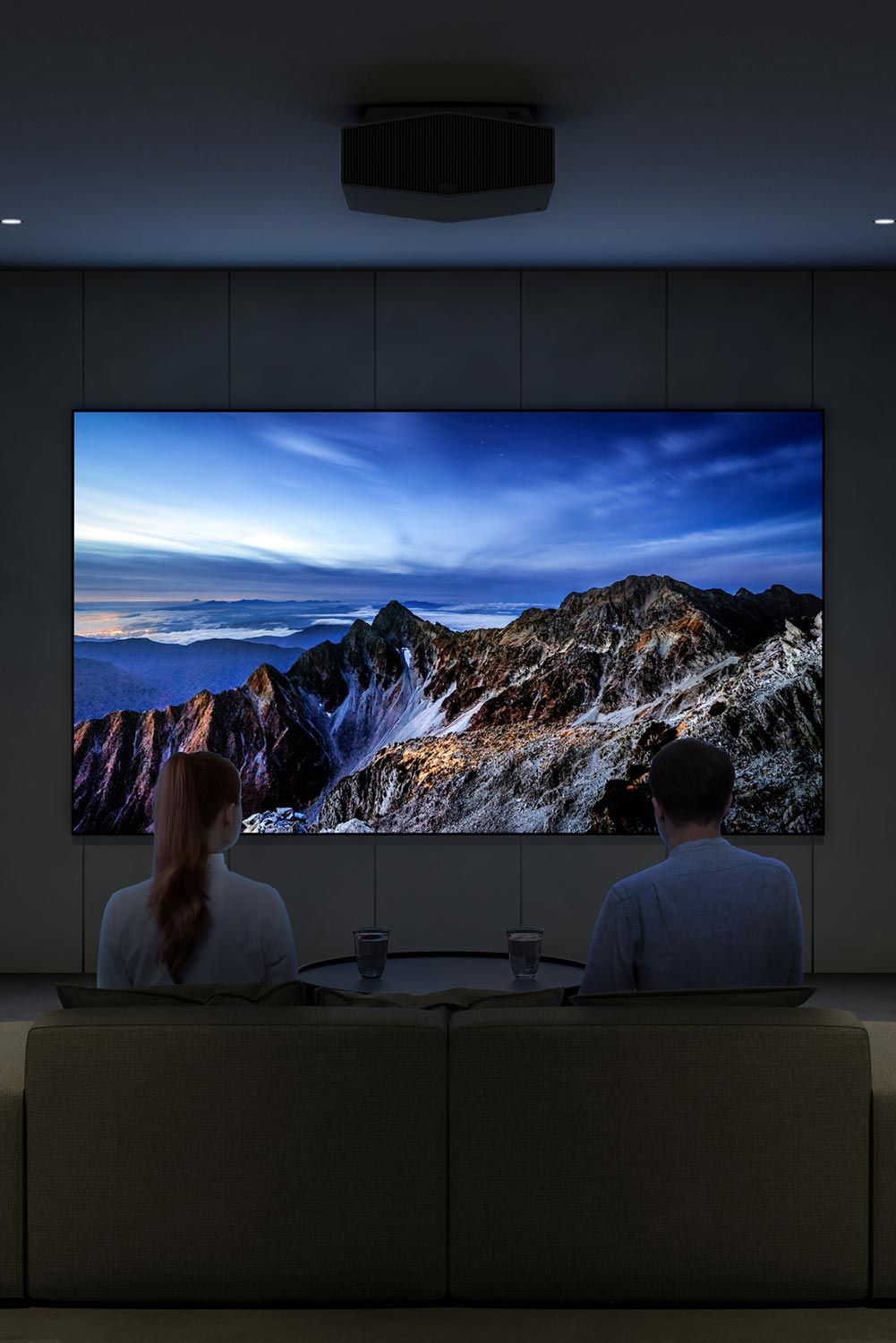 A home theater with a couple sitting on a couch, watching a large screen displaying a mountain landscape, with a ceiling-mounted projector.