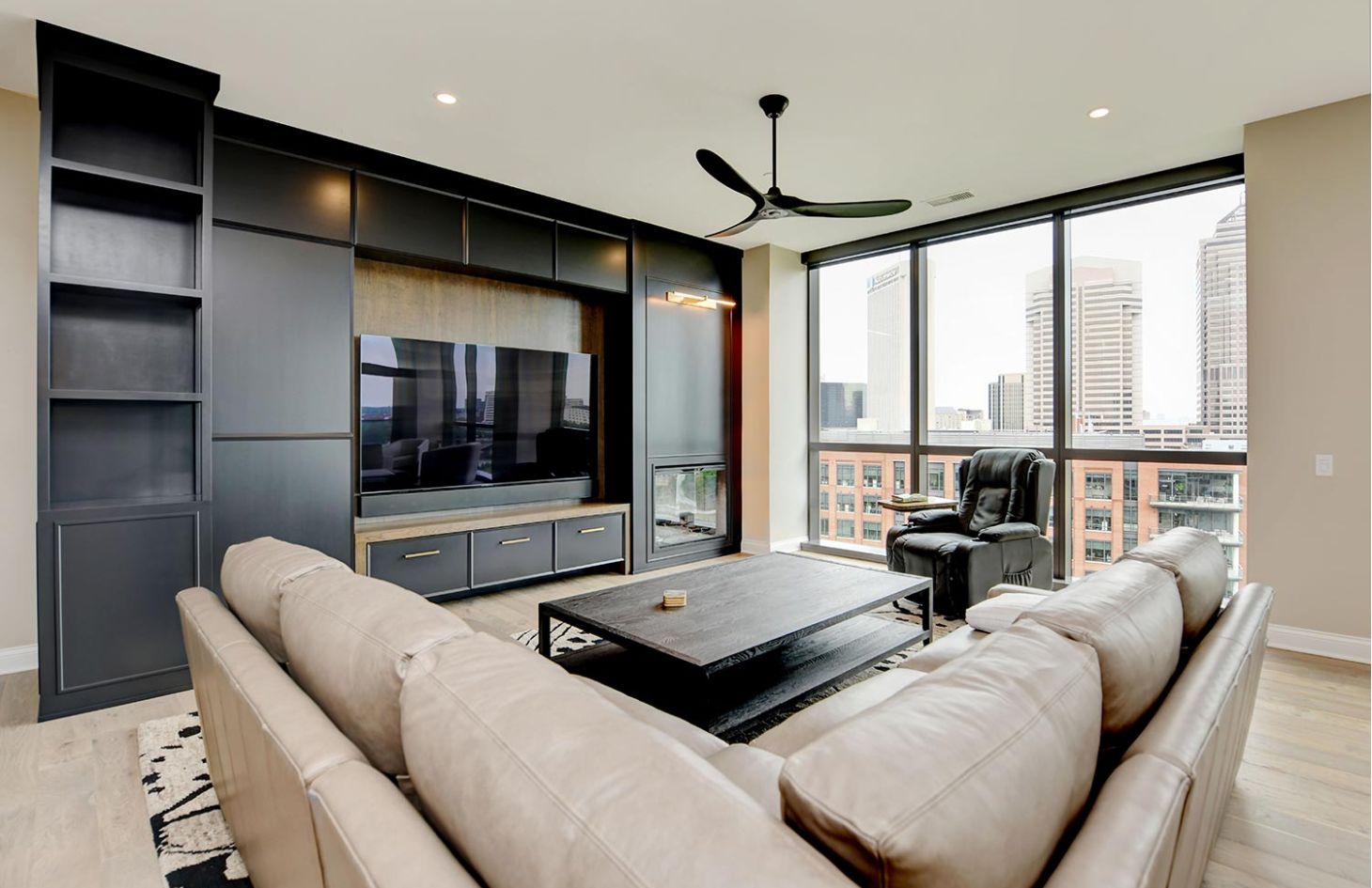 A living room with a beige sectional, black entertainment wall, and extensive city views through large windows.