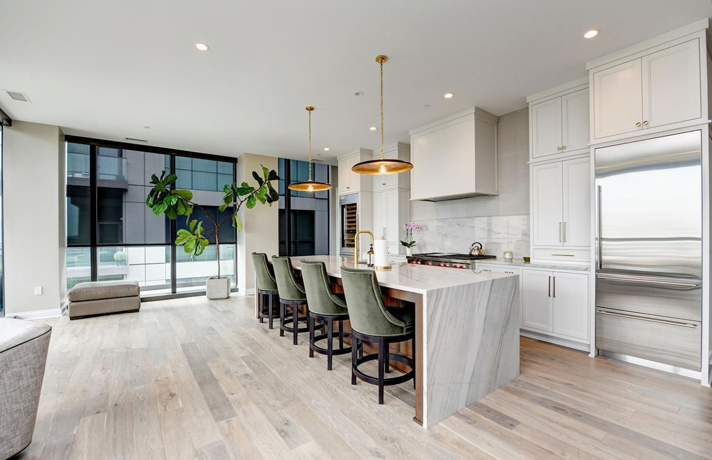 A modern kitchen with a marble island, green bar stools, and pendant lighting, highlighted by large windows.