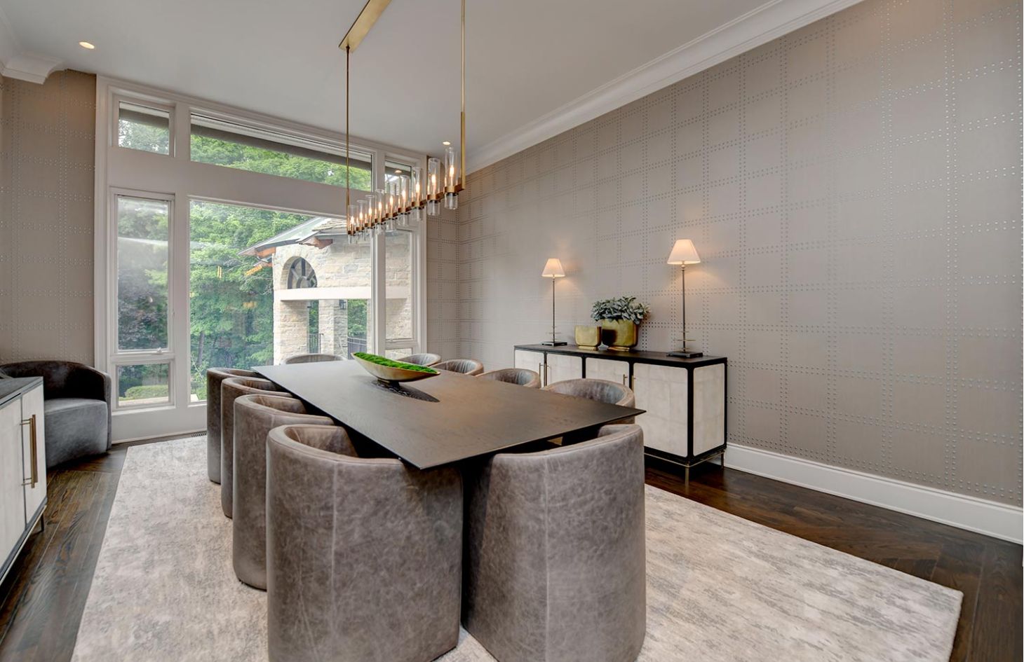 A dining room with a long table, plush seating, a contemporary chandelier, and floor-to-ceiling windows offering garden views.