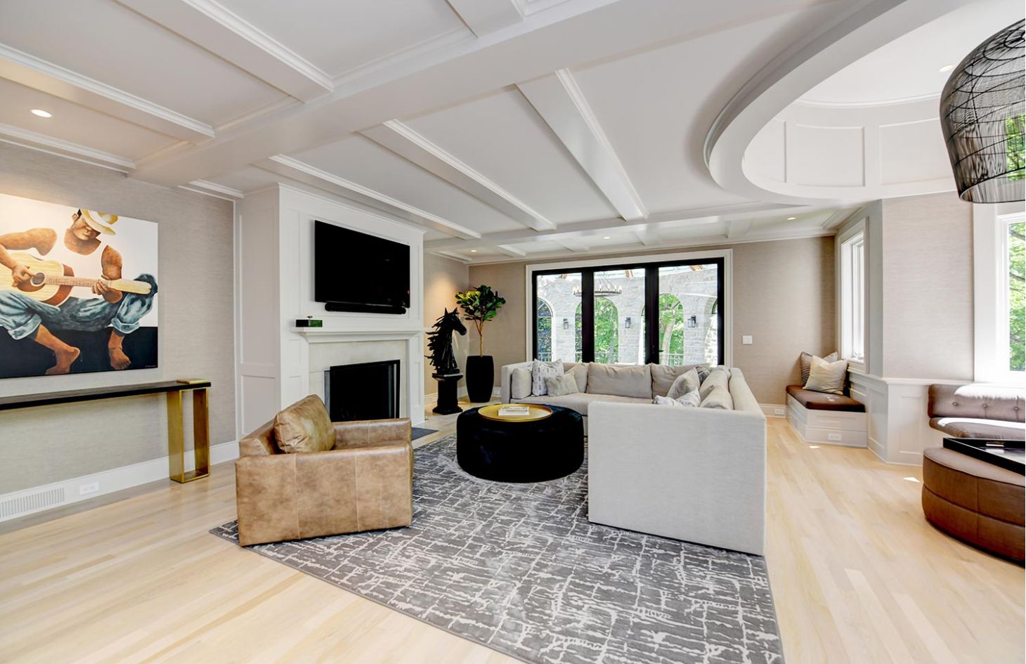 A bright living room with coffered ceilings, a large sectional, and floor-to-ceiling windows offering garden views.