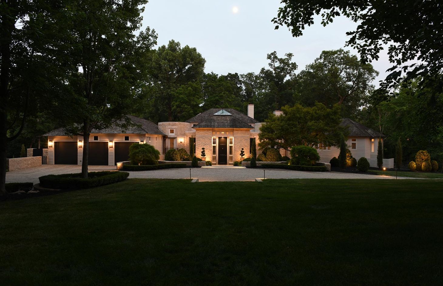 A beautifully lit home exterior at dusk with a grand entrance, manicured landscaping, and a tree-lined driveway.