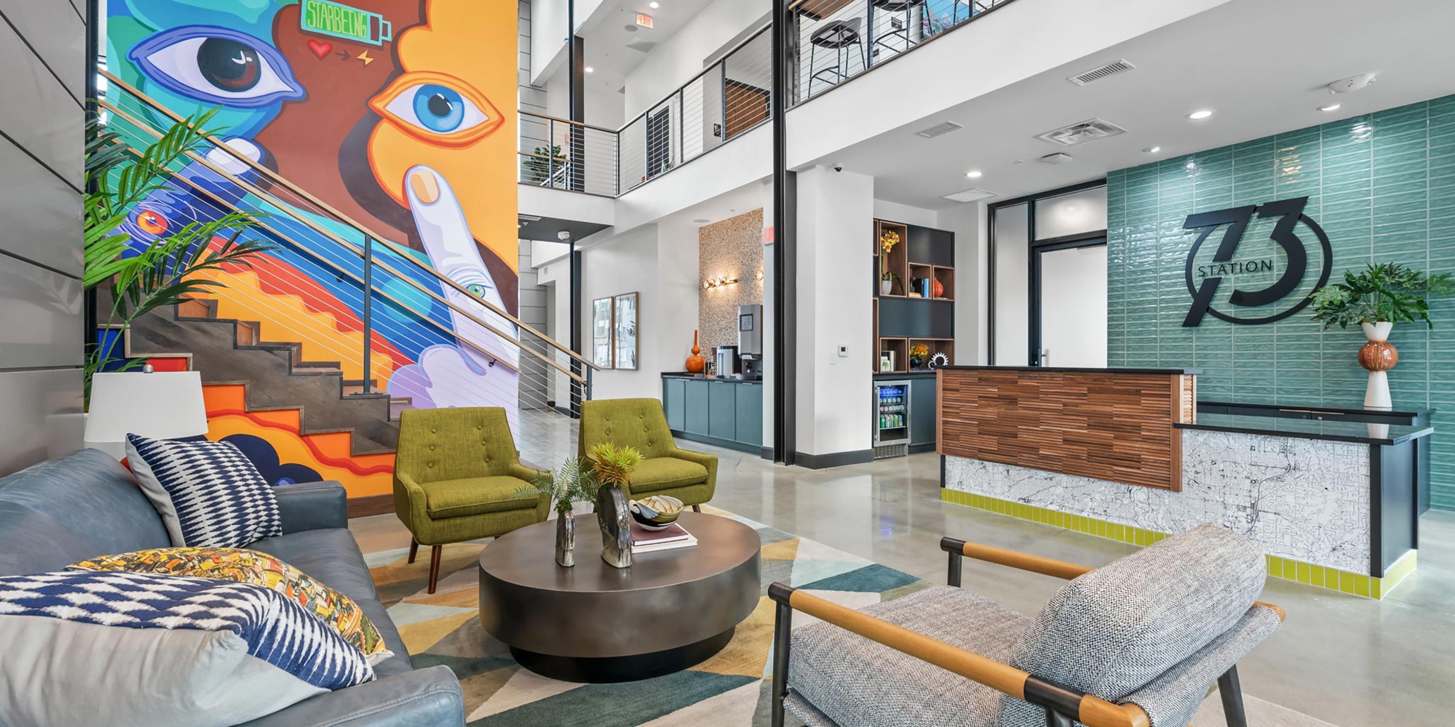 A modern lobby with vibrant wall art featuring large eyes and abstract shapes, a staircase with cable railing, green armchairs, and a reception desk with teal tiles and a geometric logo.