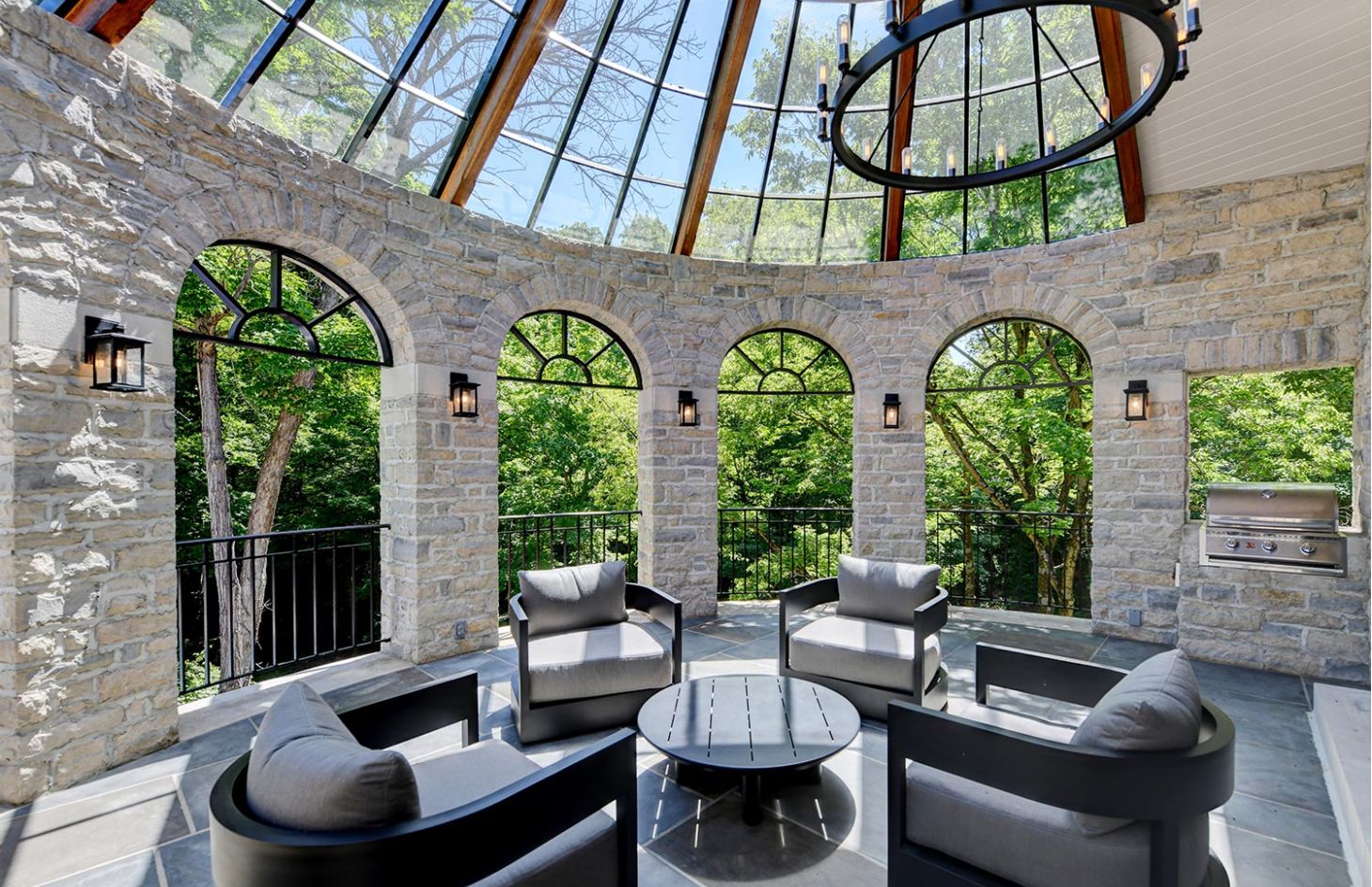 A stone patio with arched stone walls, black furniture, and large windows overlooking a wooded area.