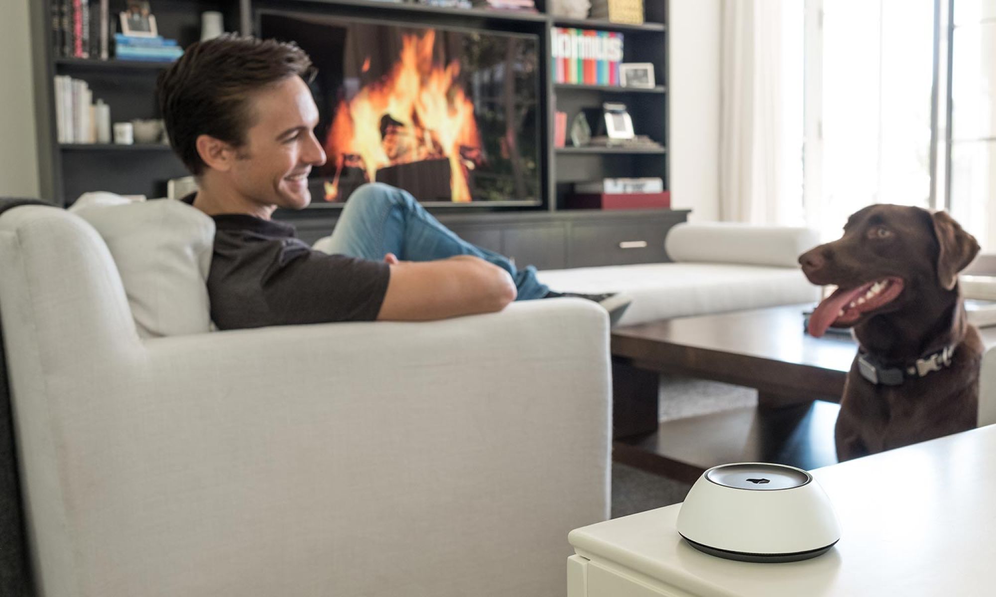 A man smiles at a dog in a cozy living room with a fireplace and a Josh AI Nano smart device on the table.