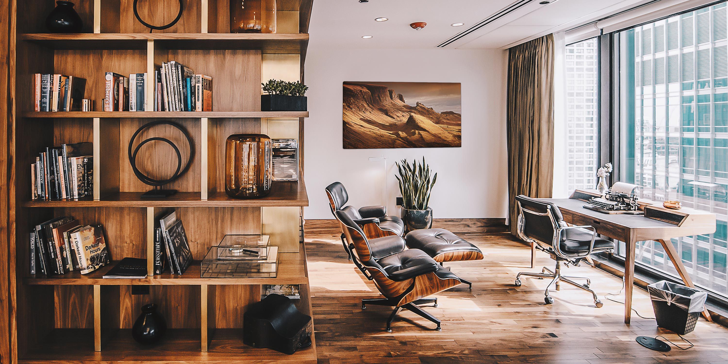 Stylish home office with a wooden bookshelf, modern furniture, and large windows offering a city view.