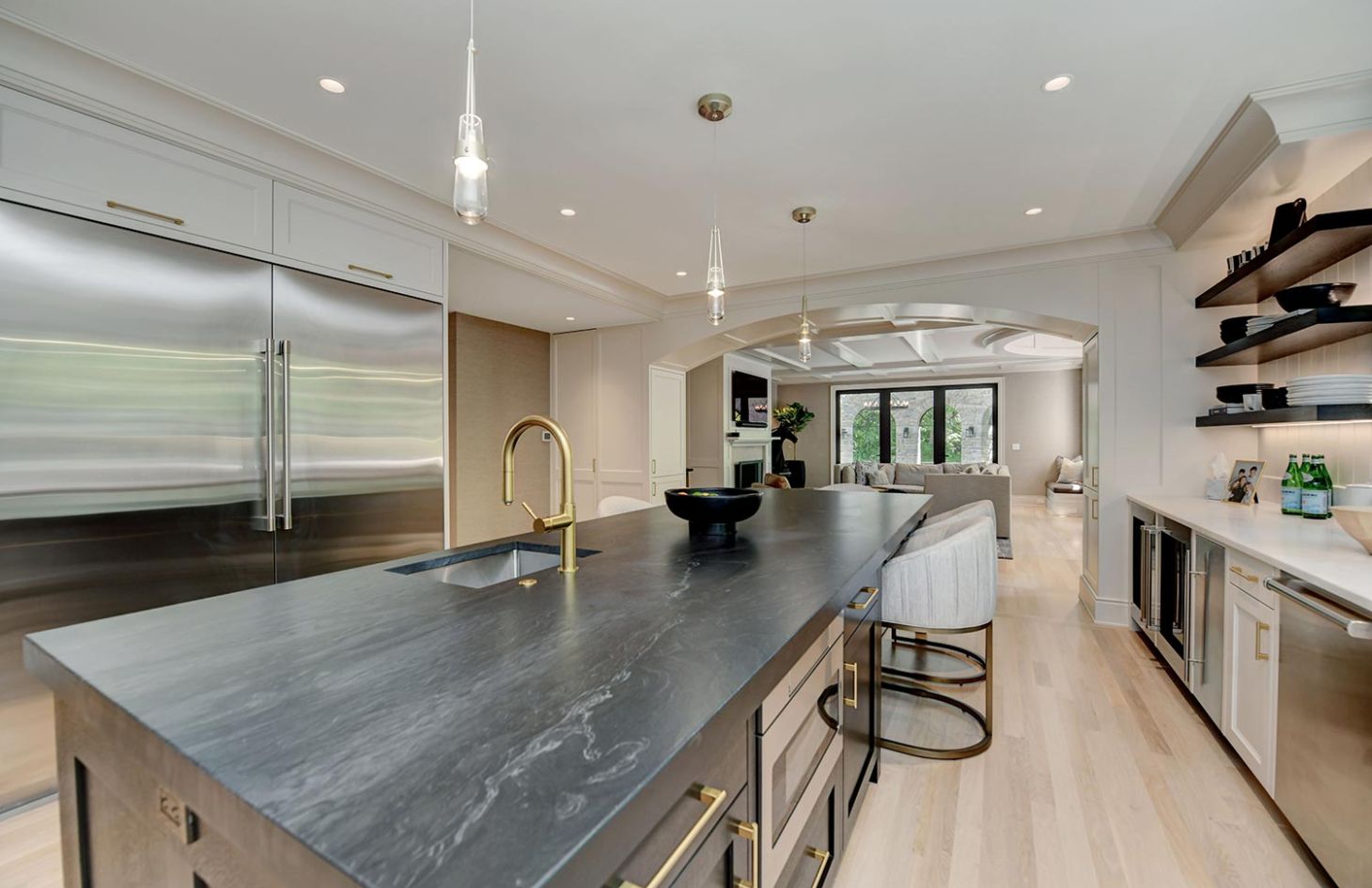 A kitchen view showcasing a sleek island with dark countertops, built-in sink, and seating, opening into a living space with large windows.