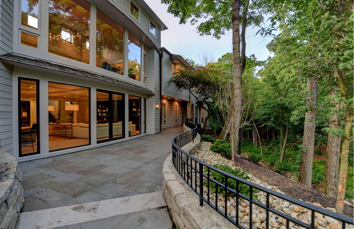 A serene back view of a home with large windows, stone patio, and a path surrounded by lush greenery.